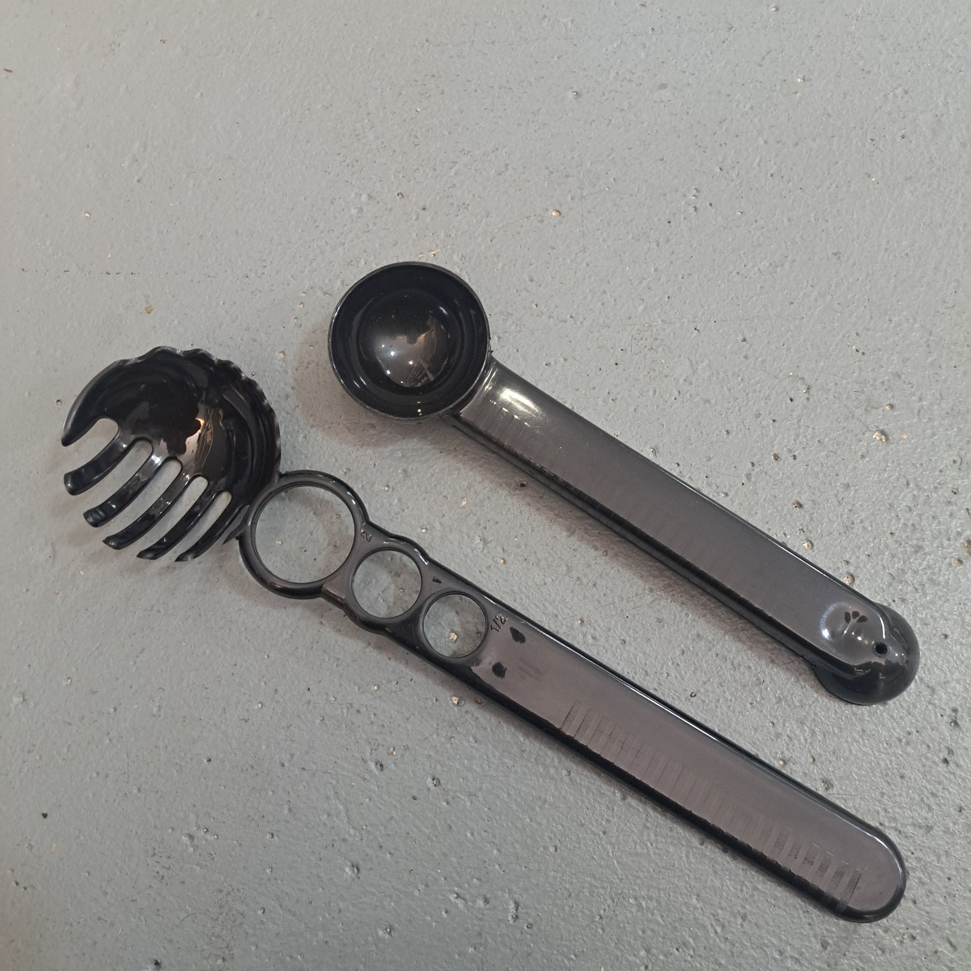 A Spaghetti Fork Scoop and an Ice Cream Scoop sit on a gray surface. The former has a toothed edge for serving pasta, while the latter features ringed handles and double-sided scooping ends.