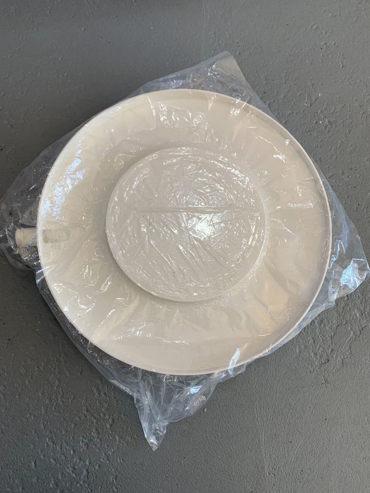 A white Drain Away Plant Drain Tray, circular and wrapped in plastic, rests on a gray surface.