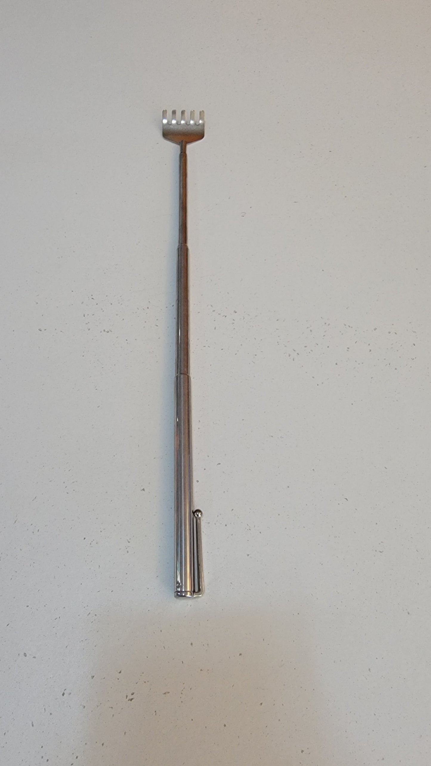 A back scratcher with a metal telescoping design and a rake-like head is extended and set on a flat surface against a plain background.