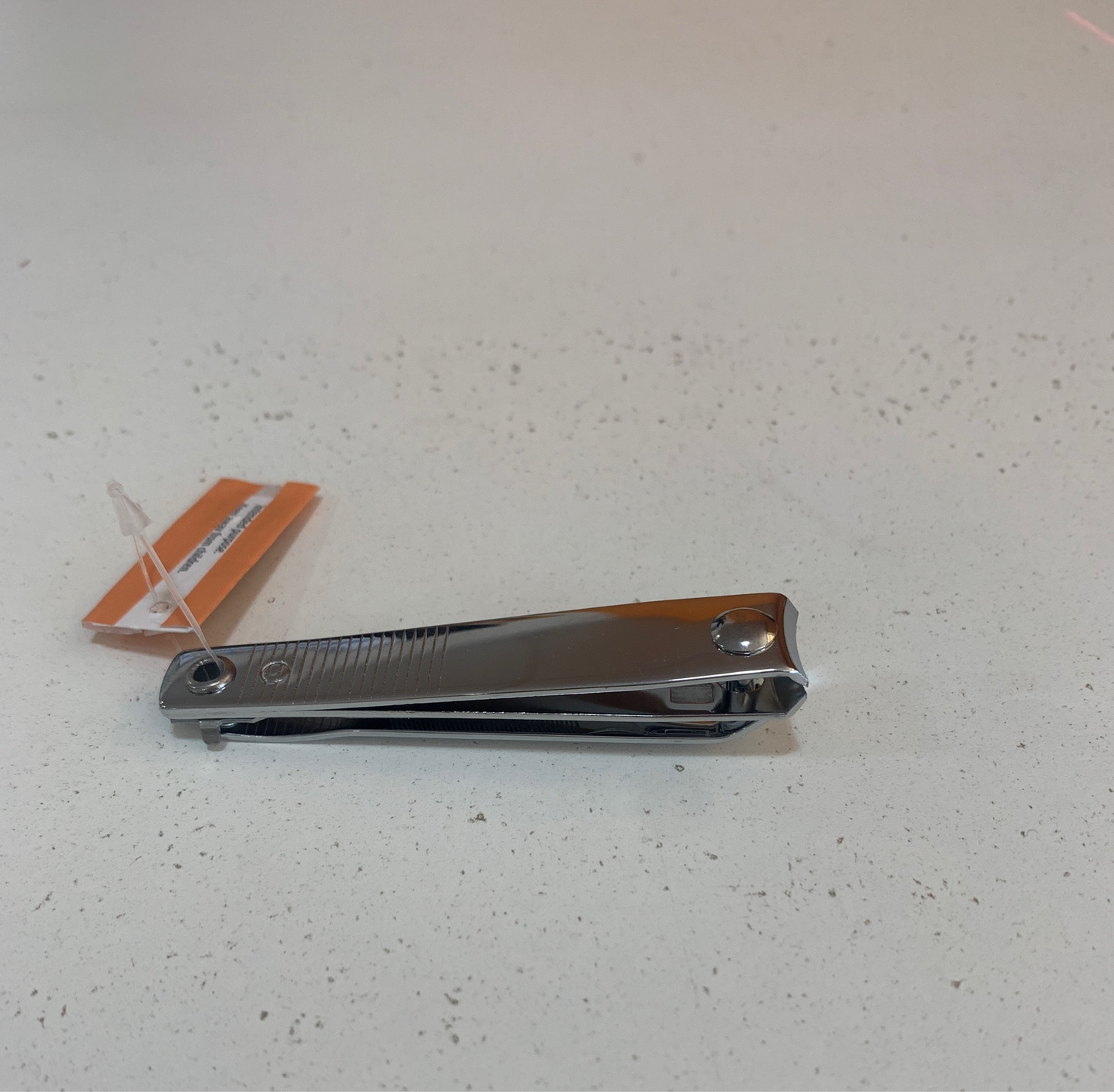 A pair of nail clippers with a small orange and white tag attached is placed on a white speckled surface.