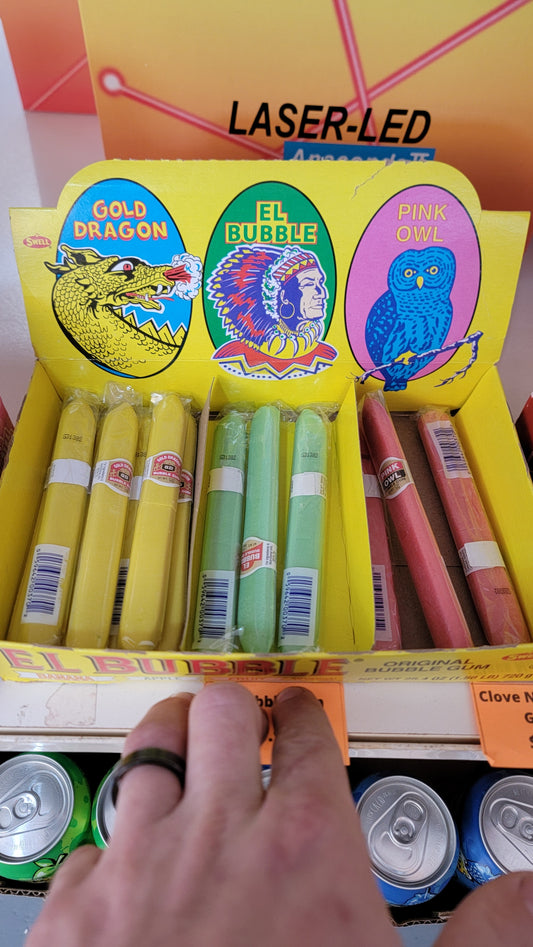 A box labeled "El Bubble Assorted Bubble Gum Cigar" is displayed on a shelf above canned drinks.