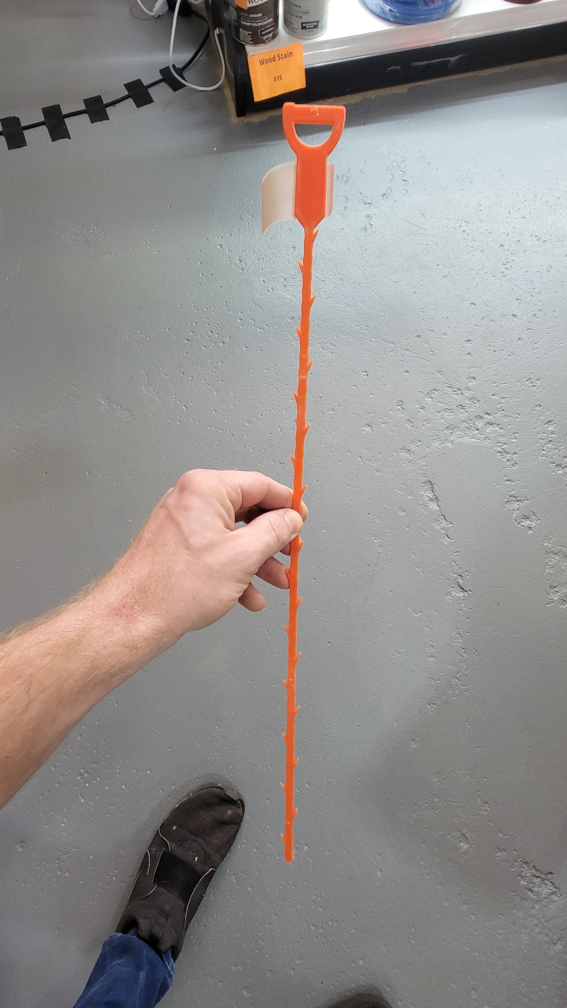 A person is holding an orange, serrated plastic drain clog removal tool with a handle at the top. The floor is gray and speckled.