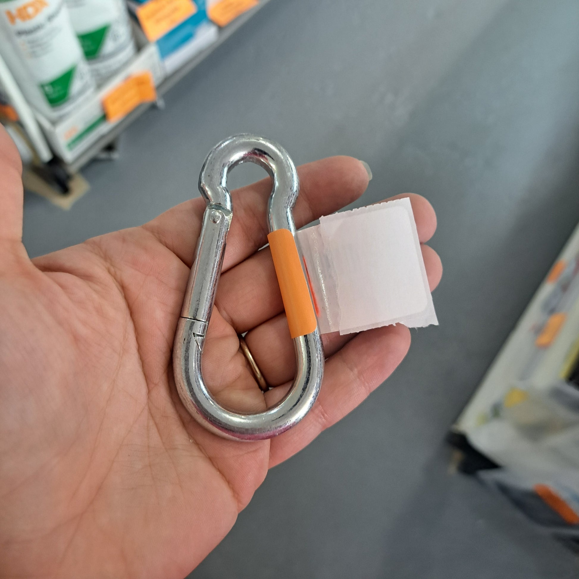 A hand holds a versatile Chrome Hook featuring an orange sleeve and a white tag, perfect for different uses. Shelves with products are visible in the background.