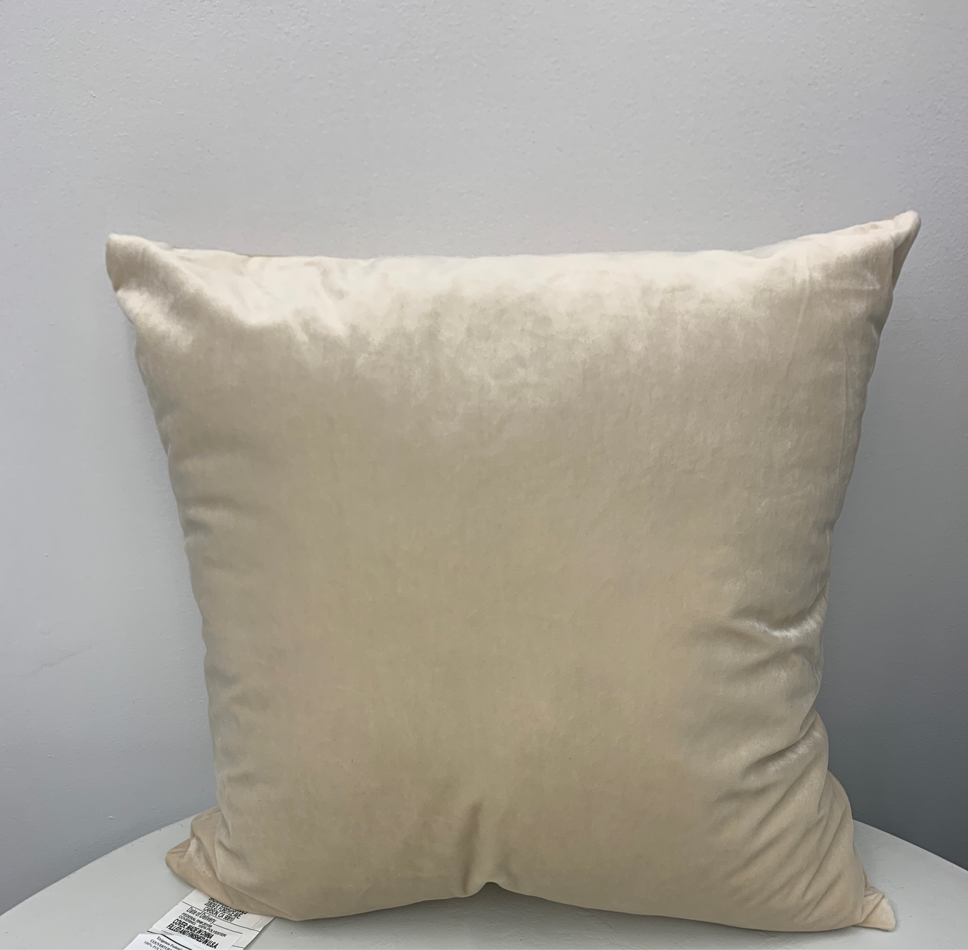 An Ivory Plush 16" x 16" Soft Faux Pillow with a smooth texture is placed against a light gray wall. A white tag with text is visible at the bottom left corner.