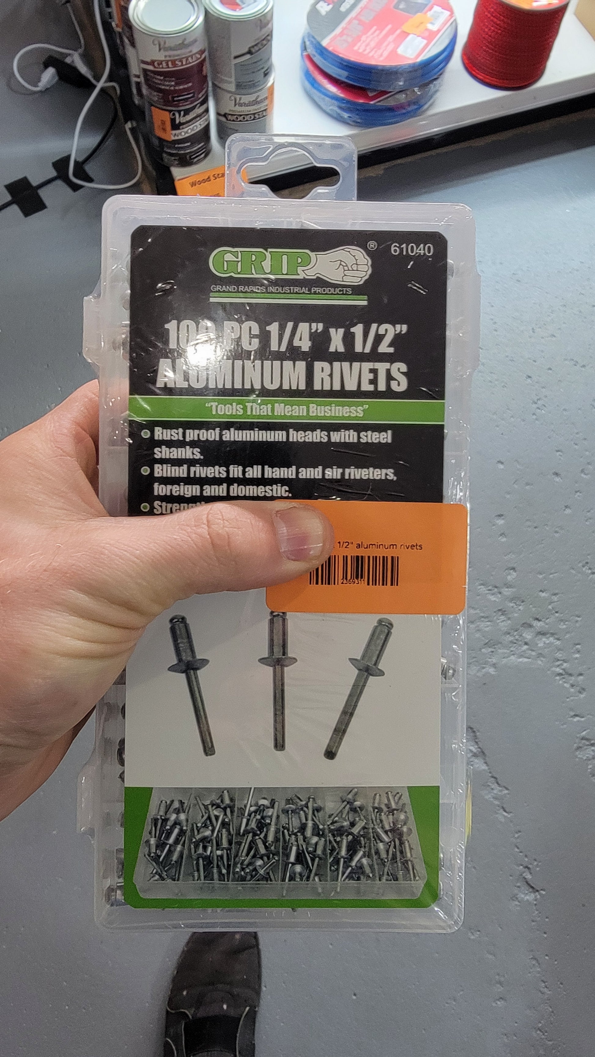 A hand holds a package of 100pc 1/4"x 1/2" aluminum rivets from GRIP, showcasing the rivets with steel shanks. Ideal for any tool box, the package features images of the rivets alongside the slogan "Tools That Mean Business.