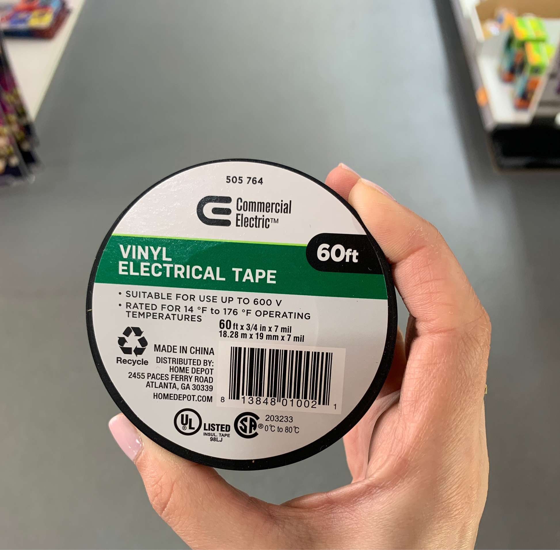 A hand holds a roll of Commercial Electric 7 Mil Vinyl Black Electrical Tape, labeled 60 feet, designed for up to 600V and varying operating temperatures, featuring recycling and safety information.