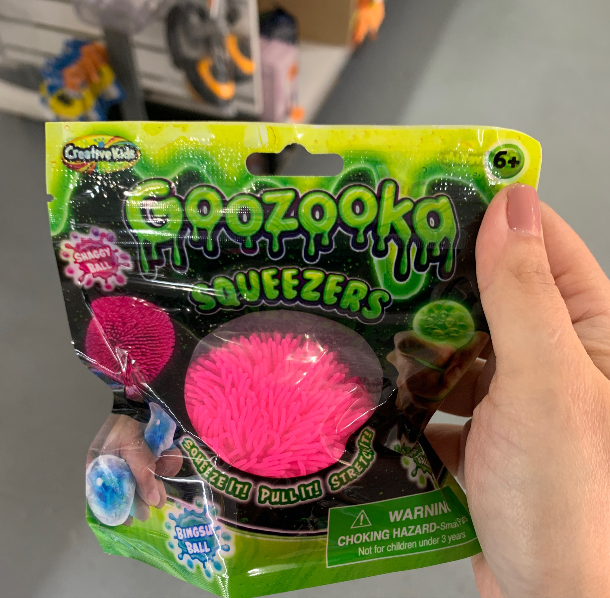 A hand holding a Goozooka Squeezers toy package, showcasing the pink color variant, labeled as suitable for children aged 6 and up and includes a choking hazard warning.