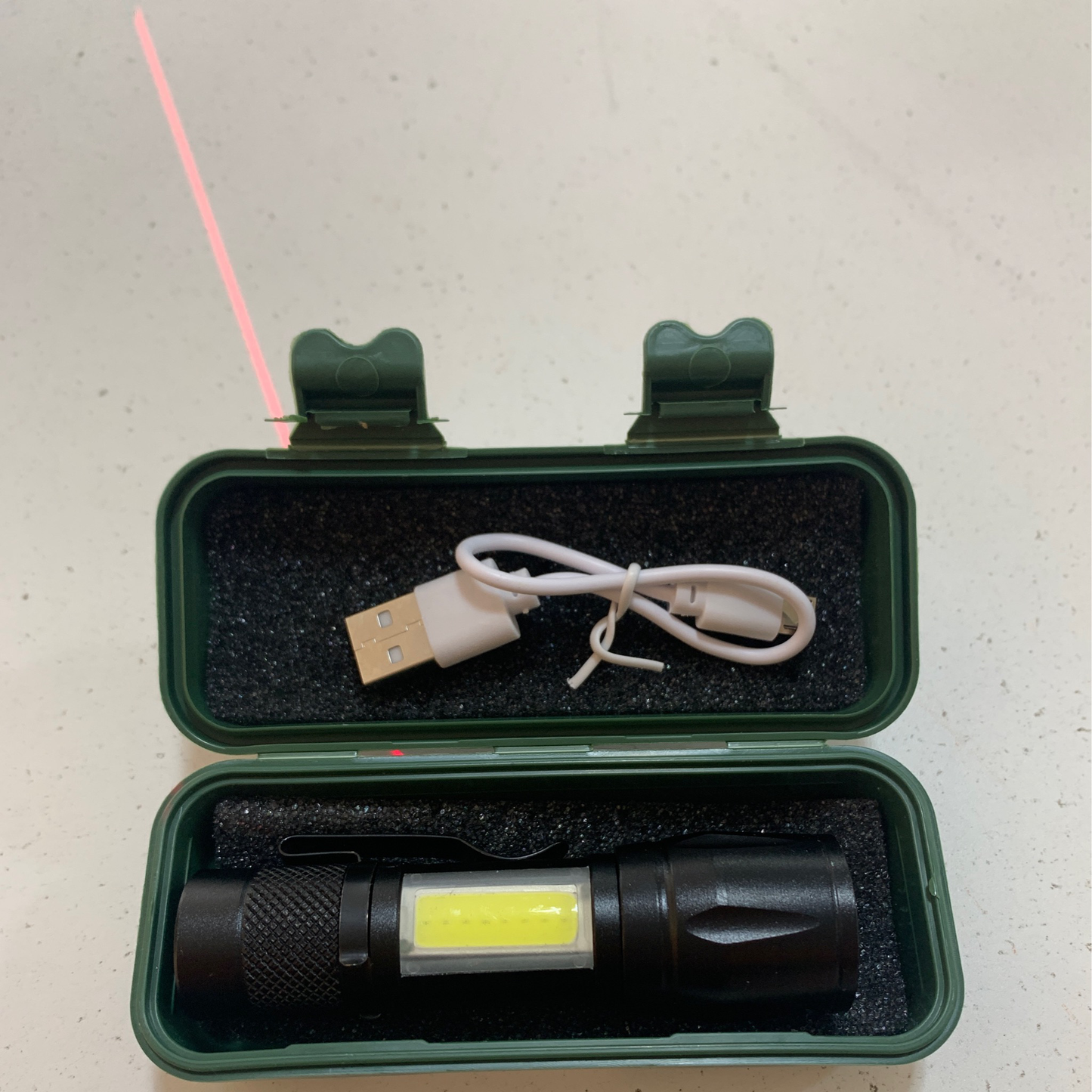 Inside the open green plastic case with foam padding, you’ll find a small black rechargeable flashlight emitting a red laser from its top, alongside a white USB charging cable.