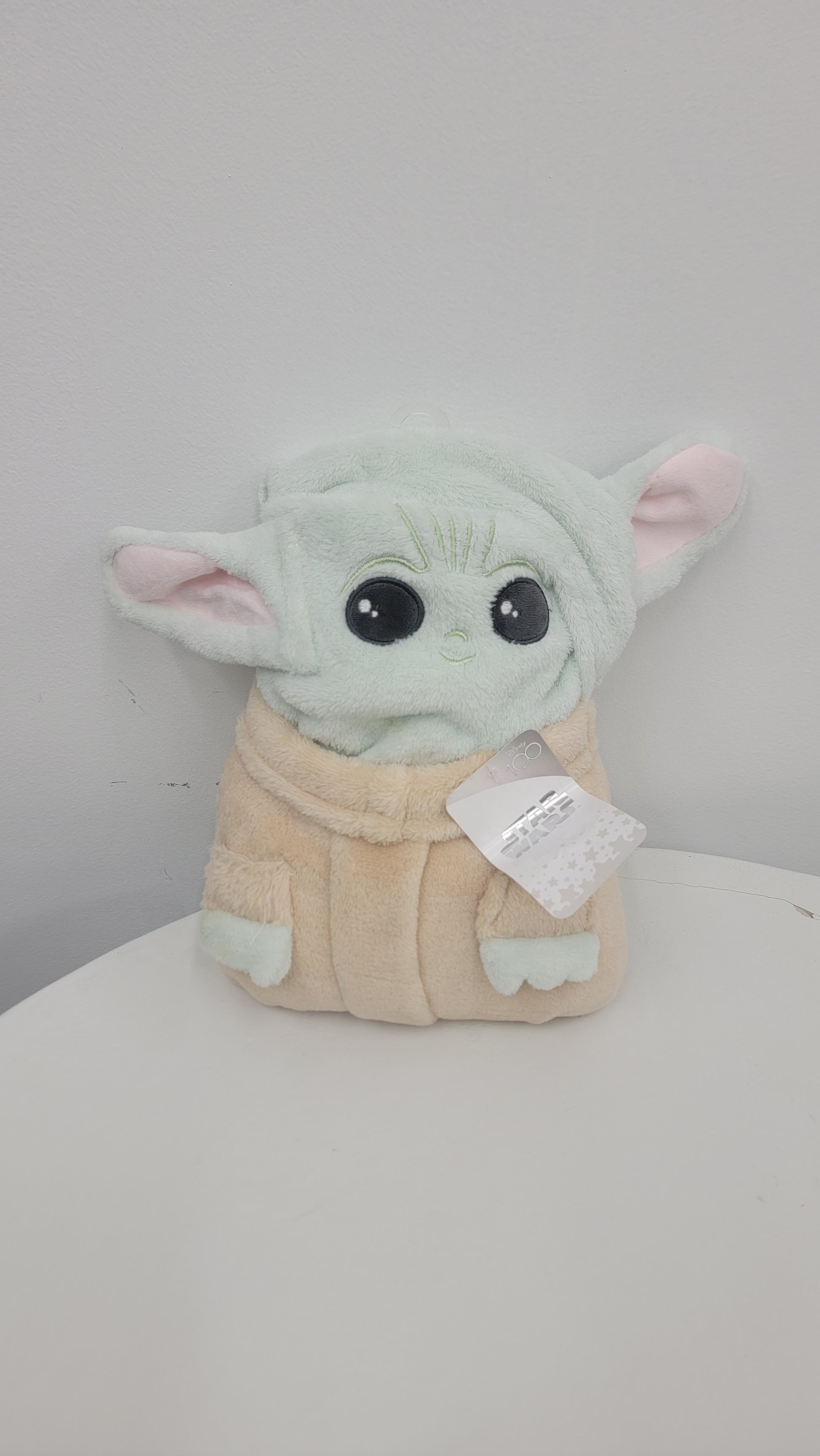 A Star Wars Character Blanket featuring The Child, a plush toy resembling a small alien creature with large eyes, pointy ears, and a beige robe, sits against a white background with its tag attached.