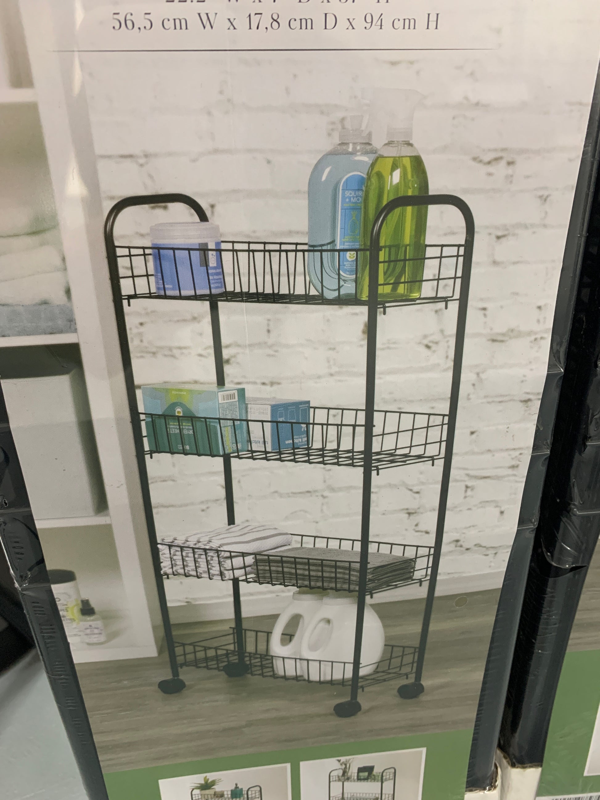 The Richards 4 Multi-Purpose Shelf Rolling Storage Cart, a four-tier metal wire shelving unit on 4 wheels, is perfect for holding cleaning supplies and toiletries. Positioned against a white brick wall backdrop, its overall dimensions of 56.5 cm W x 17.8 cm D x 94 cm H make it ideal for kitchen storage and easy transporting.