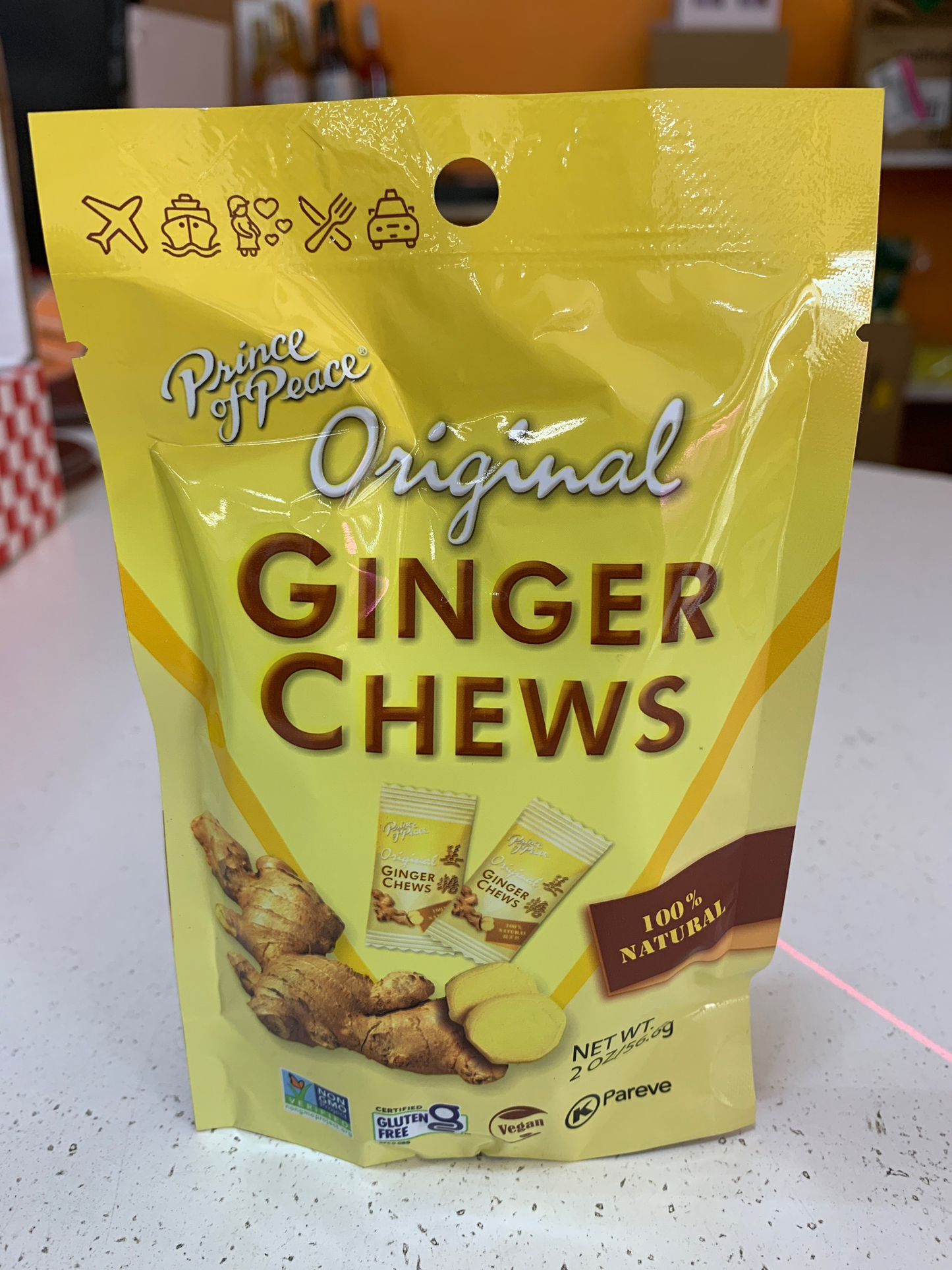 A 4 oz yellow bag of Prince Of Peace Ginger Chews, 100% natural, is displayed on a countertop. Known to help with nausea and inflammation, the packaging features images of ginger roots and individual ginger chew packets.